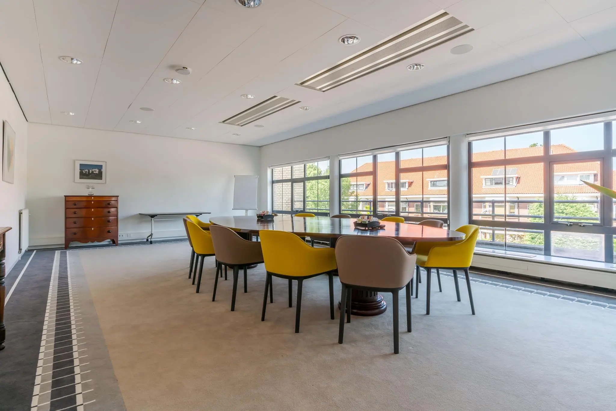 Vergaderruimte aan de Van Alkemadelaan met grote tafel, gele en beige stoelen en veel daglicht via grote ramen.